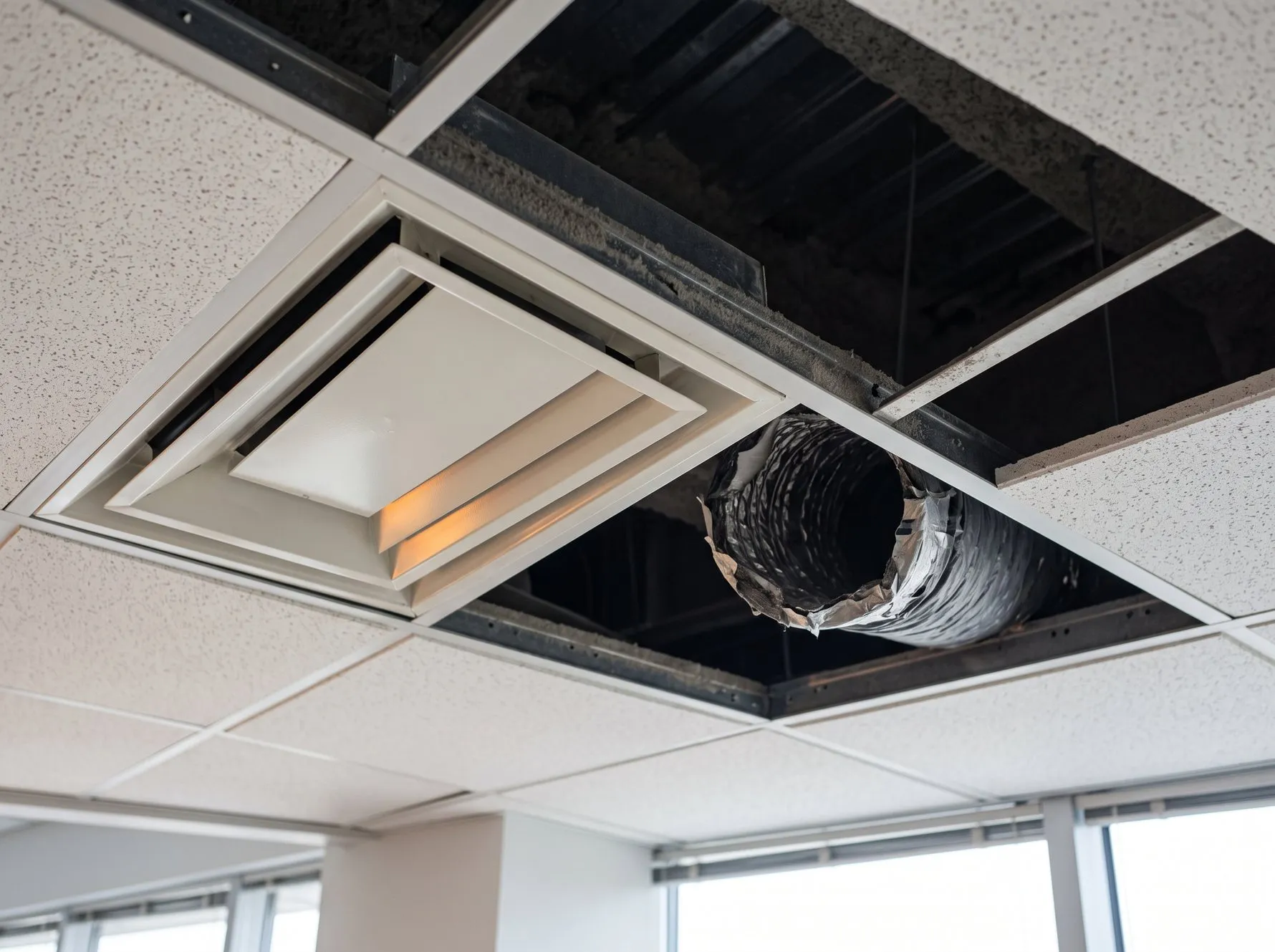 Un diffuseur d'air carré encastré dans un faux plafond de bureau, vu en contre-plongée, avec une dalle voisine déposée qui laisse voir le plénum sombre, un conduit souple isolé raccordé au réseau et la structure métallique du plancher haut.