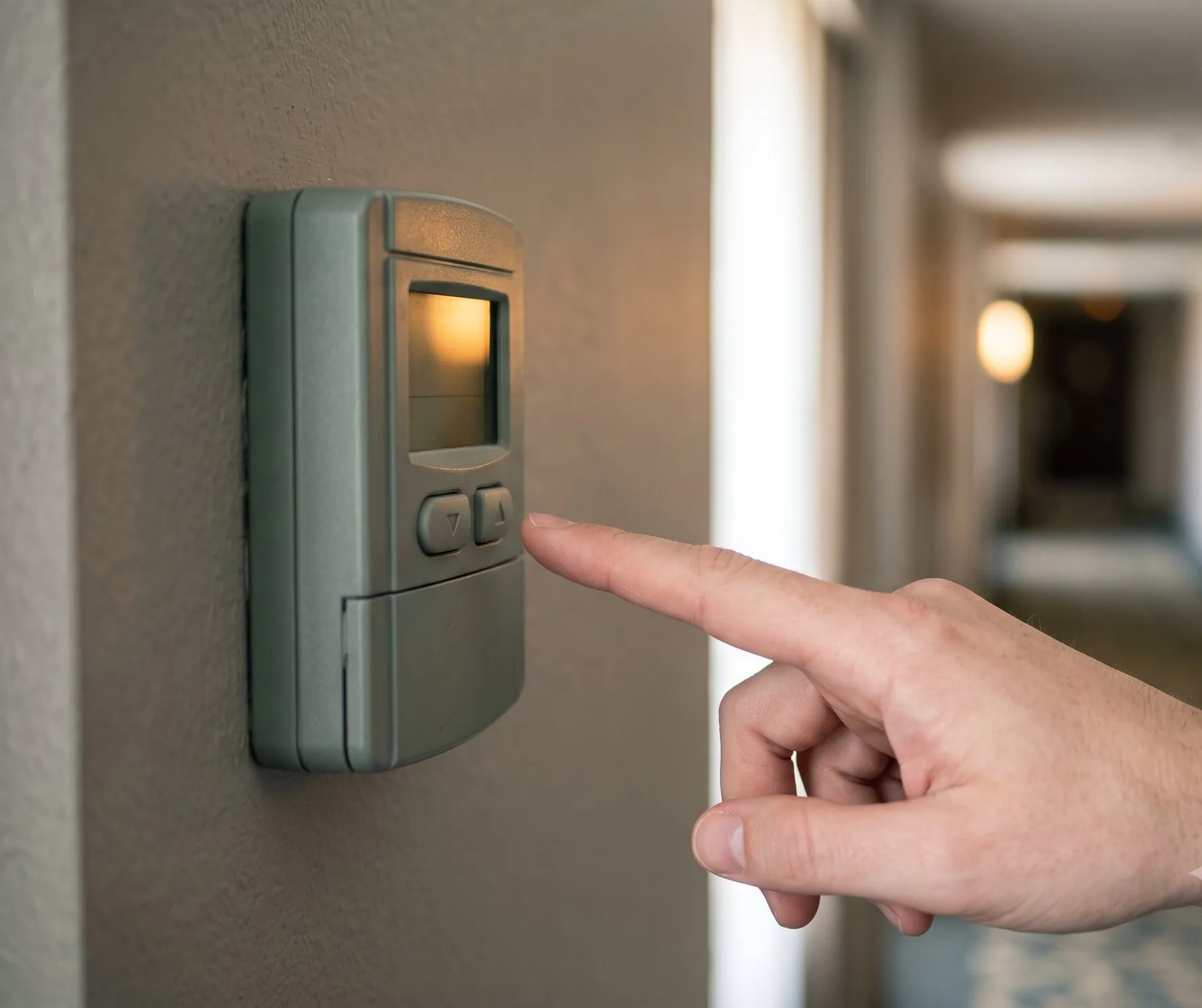 Un doigt s’approche du bouton d’un thermostat mural gris dans un couloir d’hôtel, l’écran du boîtier est éteint.
