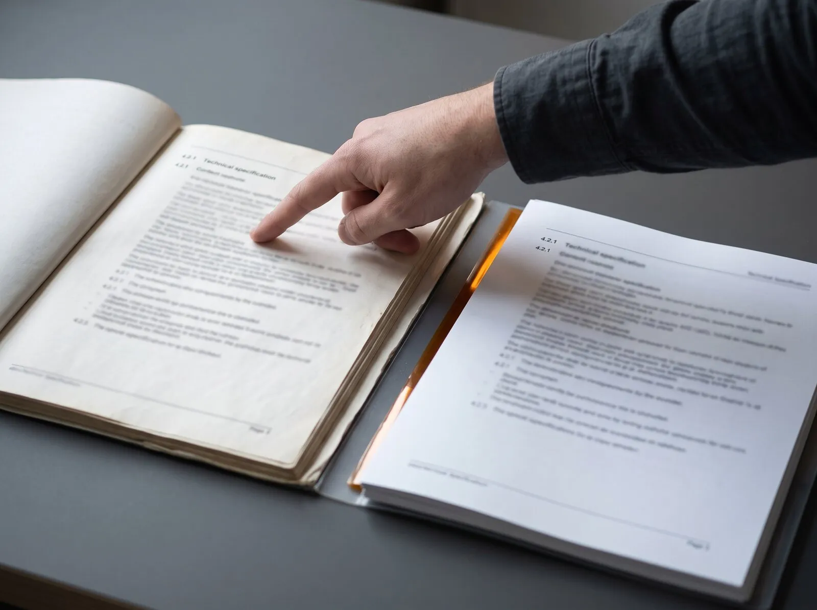 Un index pointe une ligne de texte dans un classeur ouvert et usé, à côté d'un jeu de pages neuves ouvertes à la même section, intitulée « Technical specification 4.2.1 », posées sur un bureau gris.